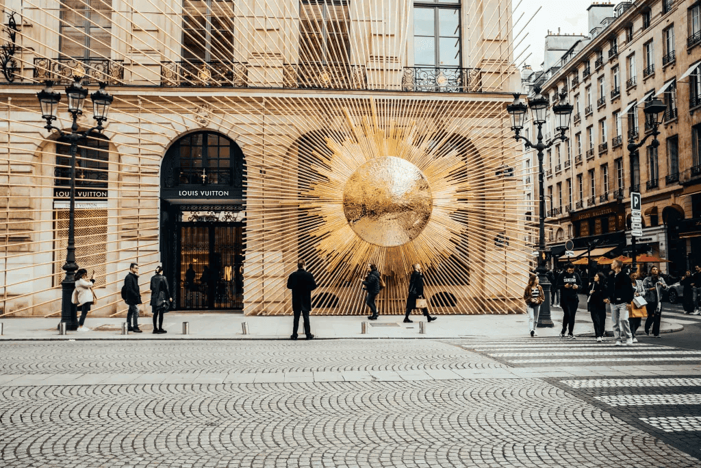 Louis Vuitton boutique facade in Paris with a large golden sun installation, shoppers walking past on a cobbled square